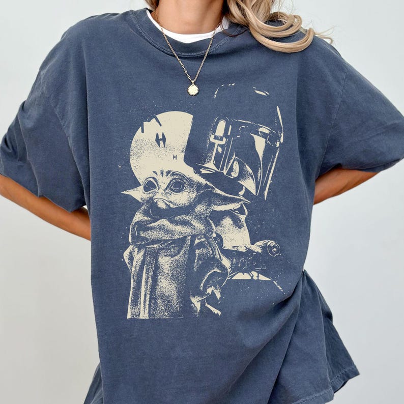 May include: Dark blue t-shirt featuring a graphic of Grogu and the Mandalorian helmet. The design is in a faded, vintage style. The shirt has a crew neck and short sleeves.