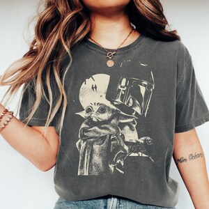 May include: Dark grey t-shirt featuring a graphic of Grogu and the Mandalorian's helmet. The design is in a vintage, distressed style. The shirt has a crew neck and short sleeves.