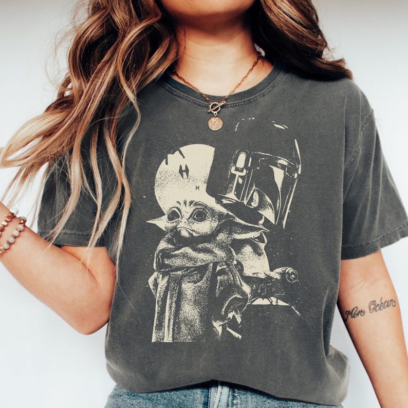 May include: Dark grey t-shirt featuring a graphic of Grogu and the Mandalorian's helmet. The design is in a vintage, distressed style. The shirt has a crew neck and short sleeves.
