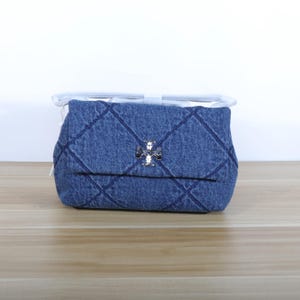 May include: A denim handbag with a quilted design and a silver clasp. The bag features a flap closure and a shoulder strap. The bag is placed on a wooden surface.