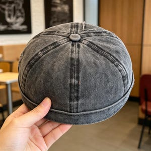 Könnte beinhalten: Graue Denim-Mütze mit einem abgenutzten, verwaschenen Aussehen. Die Mütze hat eine runde Krone, einen kleinen Schirm und ein Knopfdetail oben. Der Stoff hat sichtbare Nähte und einen verblichenen Vintage-Look.