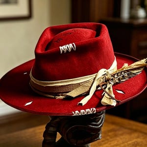 Könnte beinhalten: Ein tiefroter Fedora-Hut mit breiter Krempe. Der Hut hat ein beiges Band, eine dekorative Feder und weiße Nahtdetails. Der Hut wird auf einer dunklen Holzoberfläche präsentiert.