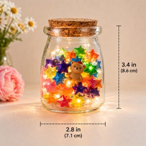 Pode incluir: Um frasco de vidro transparente com tampa de cortiça, cheio de luzes coloridas em forma de estrela e uma pequena figura de urso de pelúcia. O frasco tem 8,6 cm de altura e 7,1 cm de largura. As luzes são de várias cores.