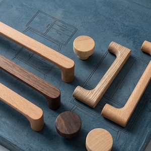 Puede incluir: Una colección de tiradores y pomos de madera para armarios, en varios tonos de marrón. La ferretería se muestra sobre una superficie azul con un diseño de plano de muebles. Los pomos son redondos y los tiradores tienen un diseño de asa curvada. Medidas en centímetros.