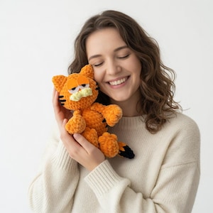 Puede incluir: Peluche Garfield naranja tejido a ganchillo, con rayas negras y hocico color crema. El peluche tiene una expresión triste con ojos naranjas. La persona lleva un jersey color crema. Artículo hecho a mano.