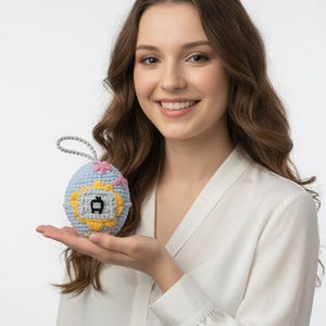 May include: A crocheted, egg-shaped ornament in light blue, yellow, pink, and grey. The ornament features a small, black and white graphic in the centre and a grey loop for hanging. The item is held in a person's hand.