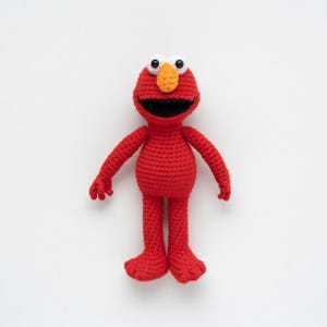 Może przedstawiać: Ręcznie robiona czerwona szydełkowa lalka z uśmiechniętą twarzą, białymi oczami i pomarańczowym nosem. Lalka ma długie ramiona i nogi i stoi wyprostowana. Lalka jest postacią z programu telewizyjnego dla dzieci.