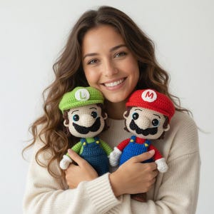 Puede incluir: Dos muñecos de ganchillo hechos a mano, uno con un gorro rojo con una "M" y el otro con un gorro verde con una "L". Ambos muñecos tienen bigotes negros, petos azules y guantes blancos. Los muñecos están inspirados en los personajes Mario y Luigi.