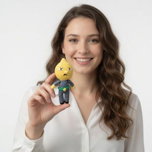 Puede incluir: Muñeca de ganchillo con forma de limón con una expresión severa. La muñeca tiene una cabeza amarilla, un cuerpo gris y un cinturón verde. La muñeca es sostenida por una persona que lleva una camisa blanca.