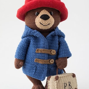 Könnte beinhalten: Gehäkelte Paddington Bär Puppe mit rotem Hut und blauem Mantel mit Knebelverschlüssen. Der Bär hält einen kleinen beigen Koffer mit den Initialen "PB" aufgestickt. Der Bär ist braun und beige.