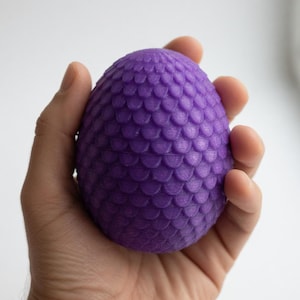 May include: A purple, egg-shaped object with a textured, scale-like pattern. The object is held in a hand against a white background. The egg appears to be made of a solid material.