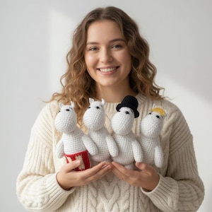 May include: Four white crocheted figures with black eyes and small arms and legs. One figure wears a red and white garment, another a black top hat, and another a yellow hair accessory. The figures are held in front of a person wearing a cream-colored sweater.