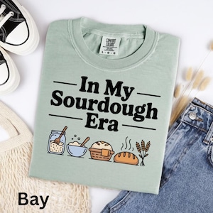 Op de afbeelding: Lichtgroen t-shirt met de zwarte tekst "In My Sourdough Era". Daaronder illustraties van bakgereedschap en brood. Het woord "Bay" staat onderaan het shirt.