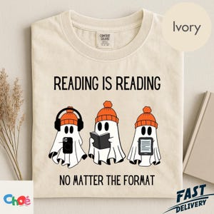 May include: An ivory t-shirt featuring the black text READING IS READING NO MATTER THE FORMAT. The graphic displays three white ghosts wearing orange beanies, each engaged in a different activity: listening to audio, reading a book, and reading an e-reader.