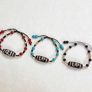 May include: Three beaded bracelets with a central black and white patterned bead. One bracelet has red beads, another has turquoise beads, and the third has black beads. All have gold-colored accents and adjustable cords.
