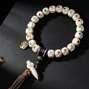 May include: A beaded bracelet featuring off-white, textured, rounded beads inlaid with small red, blue, and turquoise circular accents. The bracelet includes a gold-toned metal charm, a striped cylindrical bead, and a white pendant with a tassel.