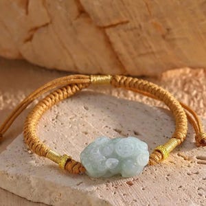 Könnte beinhalten: Ein Armband mit einem hellgrünen, geschnitzten, jadeartigen Steinanhänger in der Mitte, flankiert von zwei kleinen goldfarbenen Metallperlen. Das Band besteht aus einer geflochtenen beigen Kordel mit verstellbaren Zugbändern und goldfarbenen Fadenakzenten.