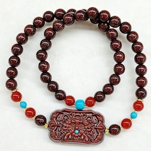 May include: Two beaded bracelets. The top bracelet is made of dark brown, round beads. The bottom bracelet has red and turquoise beads, and a carved rectangular pendant with a detailed design. Gold-colored accents are visible.