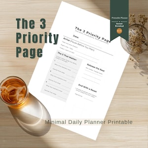 May include: A white printable planner titled "The 3 Priority Page" with sections for daily planning, including space for priorities, reflection, and a reset. The planner has a minimalist design with text in a dark green font. A glass of liquid sits on the left.