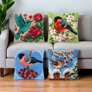 May include: Four square throw cushions featuring textured punch needle designs. One shows a teal hummingbird with red roses on a beige background, another a bullfinch on green cherry blossoms, a third a bullfinch on blue, and one a bird in snow.