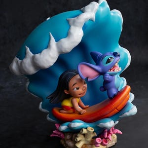 May include: A colorful figurine featuring Lilo and Stitch. Lilo is on a surfboard, and Stitch is next to her. The background is a blue wave with white accents. The base has coral and other sea elements. The figurine is a vibrant display of the characters.