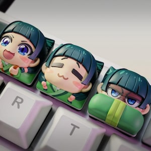 May include: Three anime-style keycaps with expressive faces, in green and tan colors, sit on a white keyboard. The keycaps feature detailed facial expressions and are designed to fit on mechanical keyboards. The keys are labeled with letters and numbers.