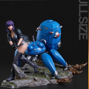 以下が含まれることがあります： 紫色の髪の女性が黒いジャケットを着て、青いロボットのようなクモ型の乗り物に乗っているフルサイズのコレクターズフィギュア。フィギュアは岩の台座に置かれています。「TURNTABLE」という言葉が左上にあります。