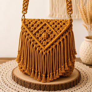 May include: A mustard yellow macrame shoulder bag featuring an intricate knotted diamond pattern on the front flap. The bag includes a thick braided strap attached with gold-toned metal rings and is finished with long, dangling fringe tassels.