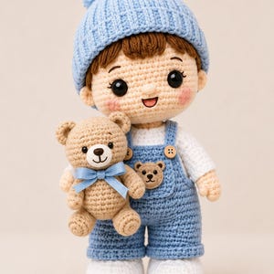 May include: A handcrafted crochet doll featuring a light blue pom-pom beanie, a white shirt, and blue overalls with a small bear patch. The doll has brown hair, rosy cheeks, and black eyes, and is holding a small tan crochet teddy bear with a blue bow.