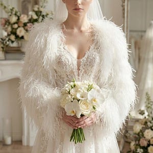May include: A white wedding dress featuring a deep V-neckline with intricate lace detailing, long sleeves adorned with soft white feathers, and a full tulle skirt. A matching pearl-embellished headband and a sheer veil are worn with the dress.