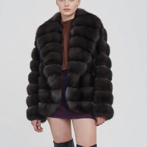 Pode incluir: Um casaco de pele castanho escuro com gola larga e design horizontal. O casaco é usado sobre um top castanho e uma saia xadrez. A modelo também usa botas pretas até ao joelho.