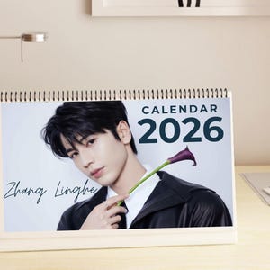 May include: A desk calendar for 2026 featuring a portrait of Zhang Linghe. The calendar has a white spiral binding and displays the word "CALENDAR" above the year. Zhang Linghe is holding a purple calla lily.