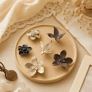 May include: A collection of six metal cabinet knobs in various floral and leaf shapes. The set includes three flower-shaped knobs in gold and dark bronze finishes, and three leaf-shaped knobs in gold and dark bronze, all featuring detailed textures.