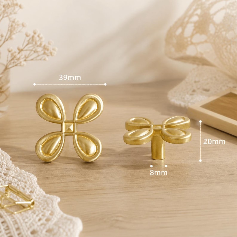 May include: Two gold-colored metal cabinet knobs with a four-petal flower design. The front view shows a width of 39mm. The side view shows a height of 20mm and a base diameter of 8mm. The knobs have a smooth, matte finish and a central square detail.