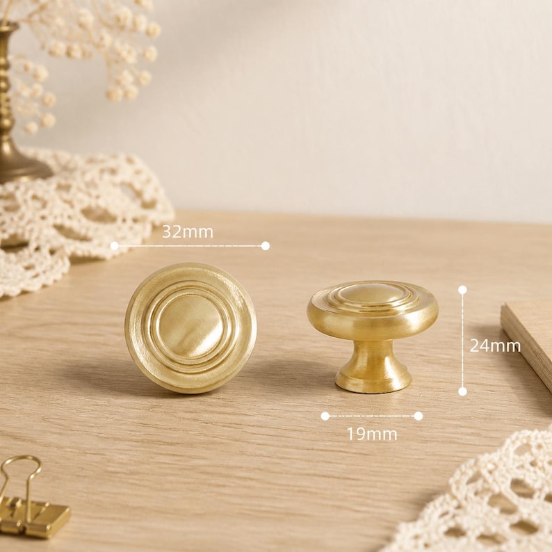 May include: Two round, brushed brass cabinet knobs with a concentric circle design. One knob is shown from the front, the other from the side. Dimensions are labeled as 32mm in diameter, 24mm in total height, and a 19mm base diameter.