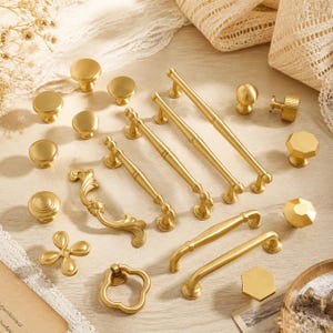 May include: A collection of gold-toned metal cabinet hardware including various drawer pulls, knobs, and handles. The set features smooth round knobs, faceted hexagonal knobs, curved ornate pulls, and straight bar handles with a brushed finish.
