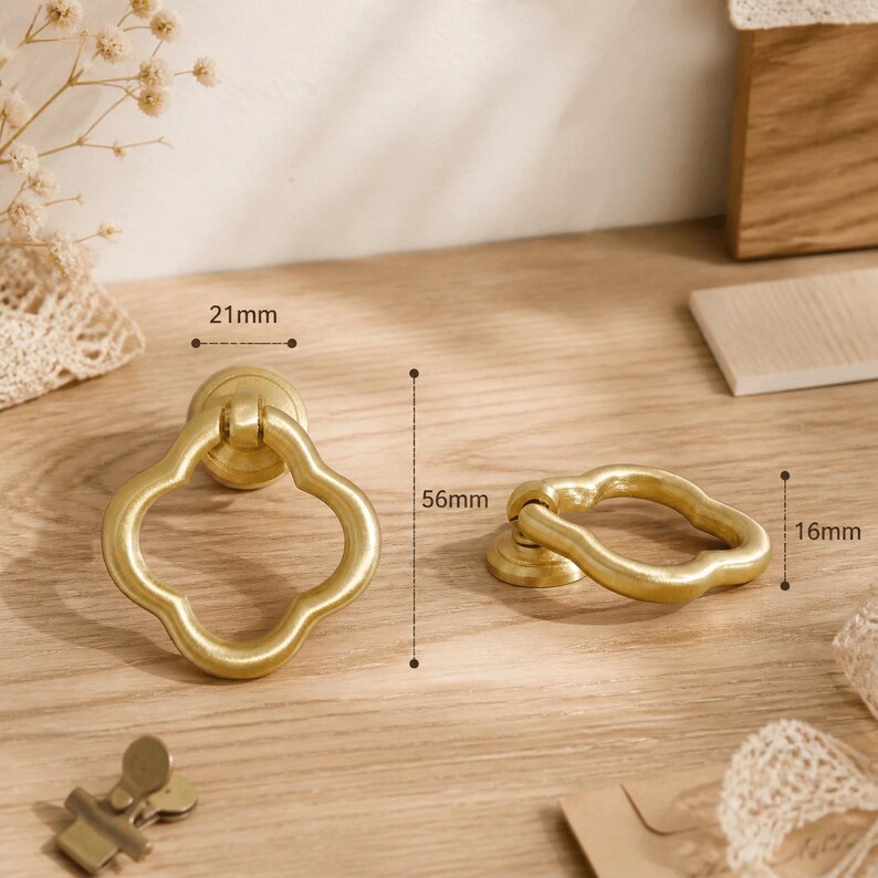 May include: Two gold-colored metal ring pulls with a clover-shaped design. The left pull measures 21mm in width and 56mm in height. The right pull measures 16mm in height. The hardware features a smooth, matte finish and a circular mounting base.