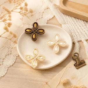 May include: Three four-petaled flower-shaped decorative buttons arranged on a small round ceramic dish. One button is finished in an antique bronze tone, while the other two feature a glossy cream-colored center with a metallic gold-toned border.