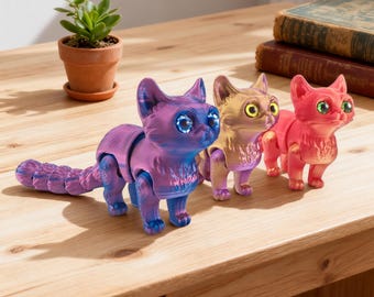 May include: Three articulated cat figurines in shades of purple, gold, and red are displayed on a wooden surface. Each cat has large, expressive eyes and a segmented tail. A small potted plant and books are in the background.