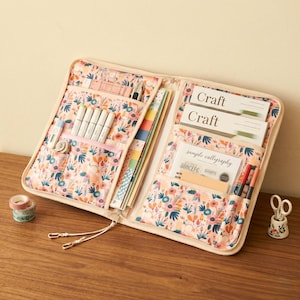 Puede incluir: Un organizador de manualidades con estampado floral con m&uacute;ltiples bolsillos y compartimentos. El organizador abierto revela bol&iacute;grafos, rotuladores, papel y otros suministros de manualidades. La tela tiene un fondo rosa con flores y hojas coloridas. El texto "Craft" es visible.