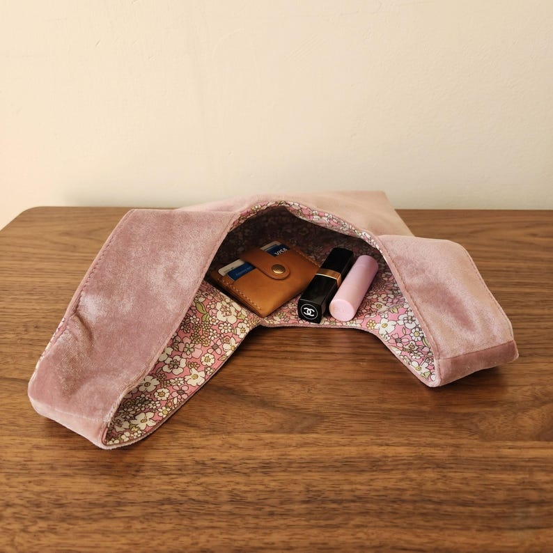 Peut inclure: Un sac hobo en velours rose poudr&eacute; avec une doublure int&eacute;rieure &agrave; motifs floraux. &Agrave; l'int&eacute;rieur du sac, on trouve un porte-cartes en cuir beige, un tube de rouge &agrave; l&egrave;vres noir avec un logo blanc et un tube de rouge &agrave; l&egrave;vres rose clair.