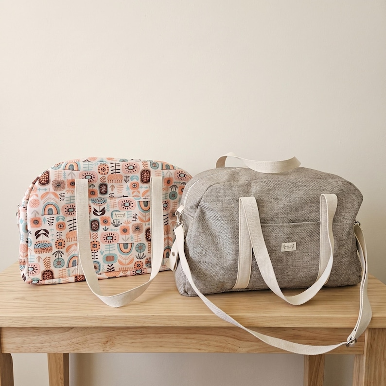 May include: Two duffel bags with white canvas handles and shoulder straps. One bag features a multi-colored pattern of abstract shapes and rainbows on a cream background. The other bag is made of a solid grey textured fabric with a small rectangular label.