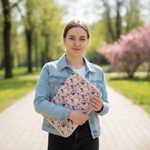 Puede incluir: Una funda para port&aacute;til con estampado floral sobre un fondo rosa claro, con flores naranjas, azules y blancas. La funda la sostiene una persona que lleva una chaqueta vaquera y pantalones negros.