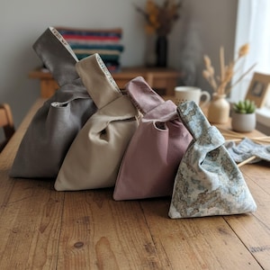 Peut inclure: Quatre sacs en tissu &agrave; n&oelig;ud dispos&eacute;s en rang&eacute;e, comprenant un sac gris, un sac beige, un sac rose poudr&eacute; et un sac &agrave; motif marbr&eacute; bleu et blanc. Chaque sac poss&egrave;de une anse unique allong&eacute;e qui se replie pour fermer le compartiment principal.