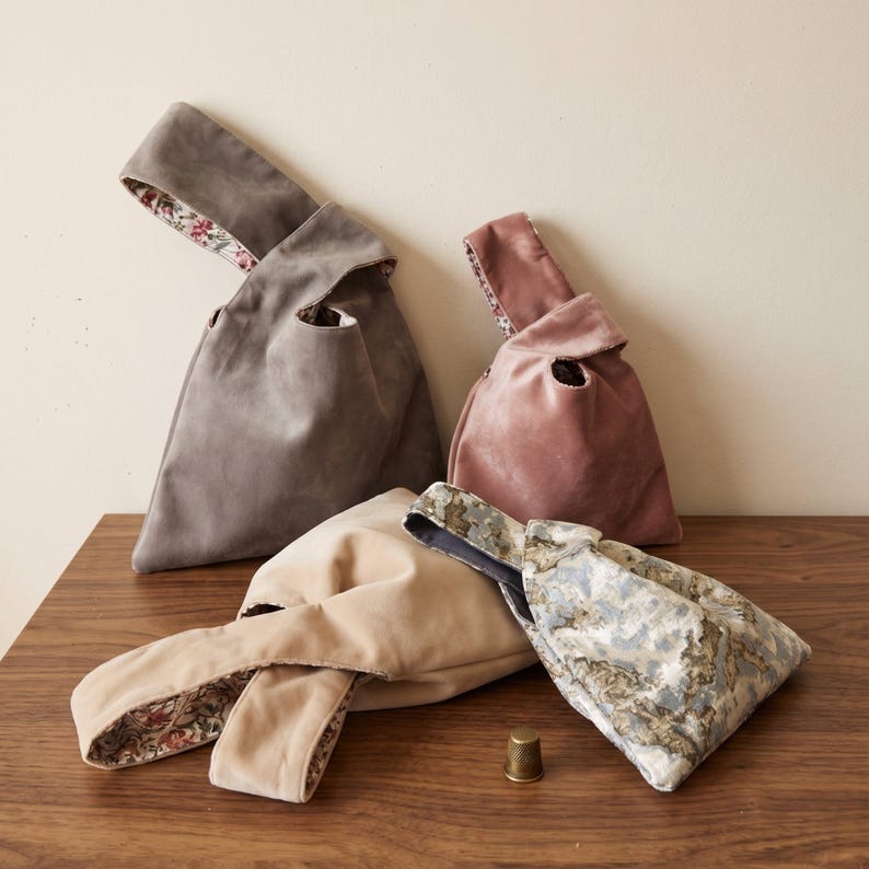 Peut inclure: Quatre petits sacs en tissu &agrave; n&oelig;ud de couleur gris, rose poudr&eacute;, beige et un motif marbr&eacute; bleu-gris. Chaque sac pr&eacute;sente une anse unique et une doublure int&eacute;rieure &agrave; motifs floraux contrastants. Un petit d&eacute; &agrave; coudre m&eacute;tallique est pos&eacute;.