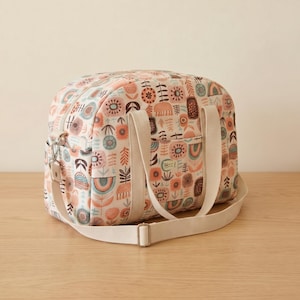 May include: A duffel bag featuring a cream-colored base with a repeating pattern of stylized flowers, leaves, and rainbows in shades of terracotta, teal, and dark brown. The bag includes two cream-colored fabric handles and an adjustable shoulder strap.