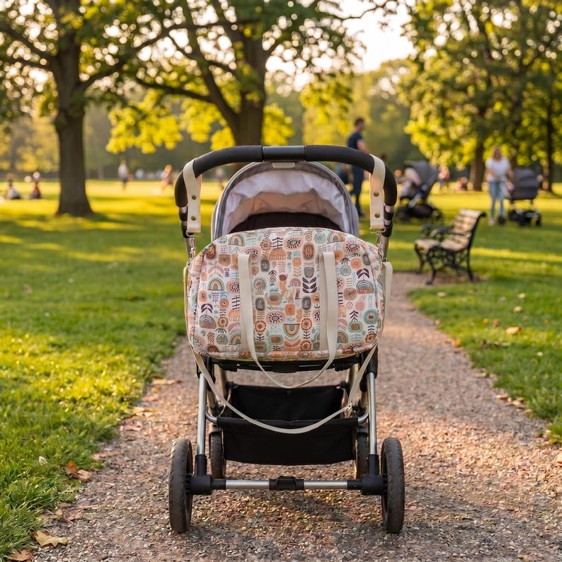 May include: A baby stroller with a silver metal frame and black wheels, featuring a cream-colored fabric bag attached to the handle. The bag displays a repeating pattern of colorful abstract shapes, including rainbows, flowers, and geometric motifs.