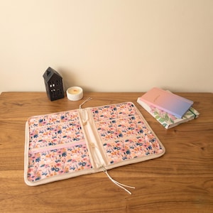 Puede incluir: Organizador abierto con estampado floral con cremallera rosa claro y bolsillos interiores. El organizador est&aacute; sobre una superficie de madera, junto a una casa decorativa negra, una vela y dos cuadernos con cubiertas florales y degradadas.