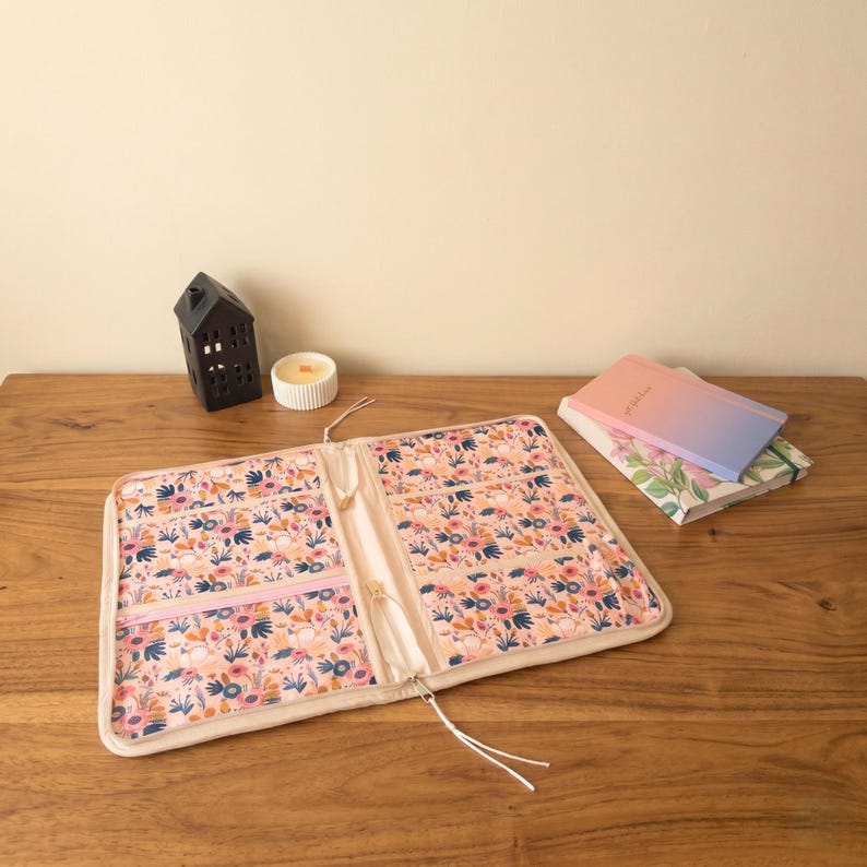Puede incluir: Organizador abierto con estampado floral con cremallera rosa claro y bolsillos interiores. El organizador est&aacute; sobre una superficie de madera, junto a una casa decorativa negra, una vela y dos cuadernos con cubiertas florales y degradadas.