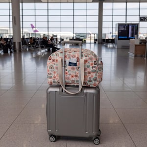 May include: A silver hard-shell carry-on suitcase with a patterned fabric travel bag resting on top. The travel bag features a colorful geometric and floral print, cream-colored handles, and an exterior pocket holding a passport and boarding pass.
